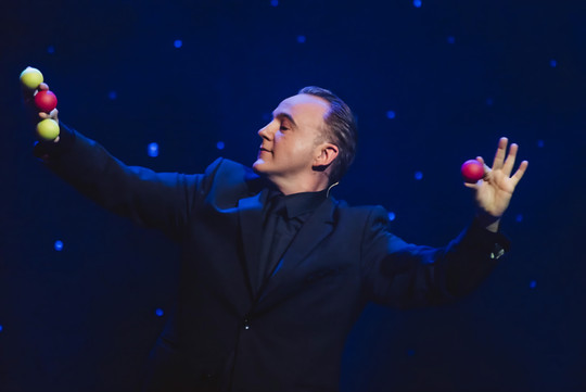 Norbert Ferré performing sleight of hand with red and yellow balls, his arms outstretched on stage.