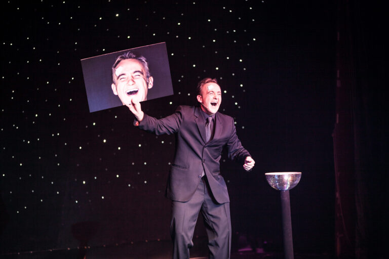 Norbert Ferré laughing on stage while holding a large smiling portrait.